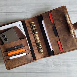 May include: A brown leather planner with a brass ring binder, a pen, and a phone in the pocket. The planner has multiple pockets and slots for cards and other items.