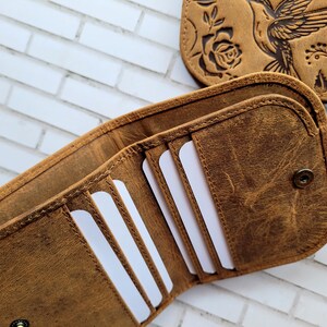Puede incluir: Una cartera plegable de cuero marr&oacute;n con cierre de broche, que cuenta con m&uacute;ltiples ranuras para tarjetas en el interior y un panel exterior a juego grabado con un dise&ntilde;o de colibr&iacute; y rosa. La cartera se muestra abierta.