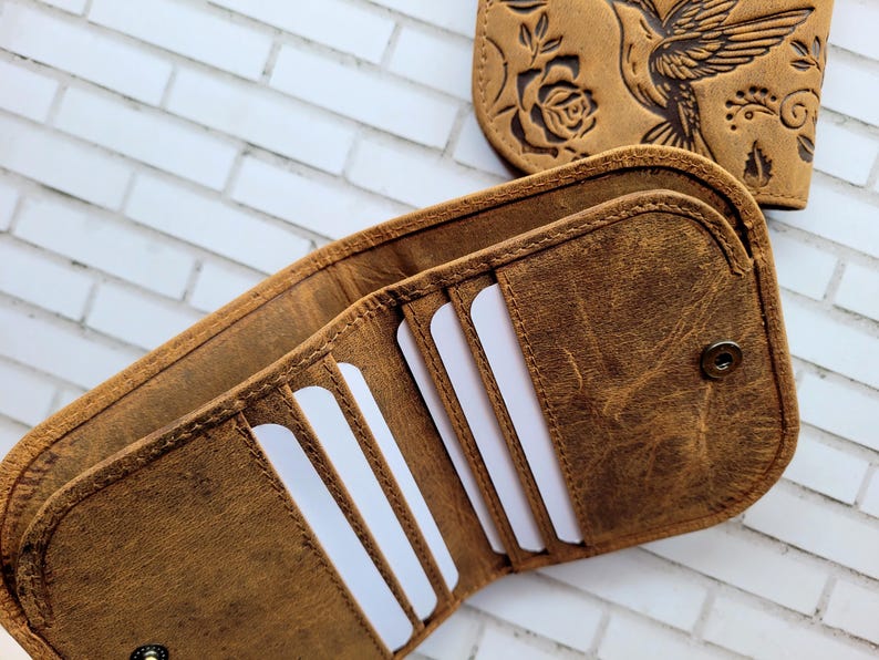 Puede incluir: Una cartera plegable de cuero marr&oacute;n con cierre de broche, que cuenta con m&uacute;ltiples ranuras para tarjetas en el interior y un panel exterior a juego grabado con un dise&ntilde;o de colibr&iacute; y rosa. La cartera se muestra abierta.
