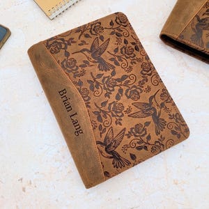 May include: Brown leather journal with a hummingbird and floral design. The journal cover features the name "Brian Lang" and is surrounded by a detailed pattern of hummingbirds and roses. The journal is rectangular and has a worn, vintage look.