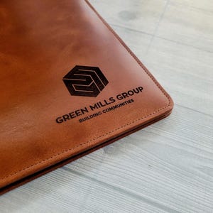 May include: A brown leather folder with a stitched edge. The folder has a logo embossed on the front that says "Green Mills Group Building Communities".