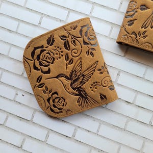 Puede incluir: Cartera de cuero color canela con dise&ntilde;o de colibr&iacute; y rosas. La cartera presenta un patr&oacute;n en relieve detallado de un colibr&iacute; en vuelo, rodeado de rosas y hojas. La cartera es de color marr&oacute;n claro.