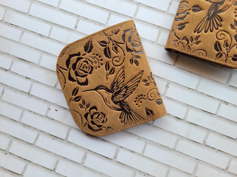 Puede incluir: Cartera de cuero color canela con dise&ntilde;o de colibr&iacute; y rosas. La cartera presenta un patr&oacute;n en relieve detallado de un colibr&iacute; en vuelo, rodeado de rosas y hojas. La cartera es de color marr&oacute;n claro.