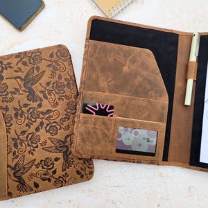 May include: Brown leather portfolio with a hummingbird and rose design. The portfolio is open, revealing pockets for cards, a pen holder, and a notepad with a sketch. The cover has a distressed finish.