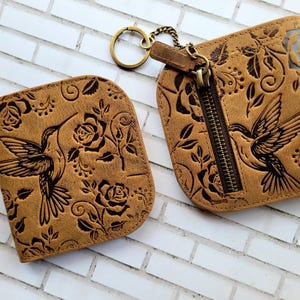 Puede incluir: Dos carteras de cuero marr&oacute;n con dise&ntilde;os de colibr&iacute;es y rosas. Una cartera tiene cremallera y llavero. La otra es un tarjetero. Las carteras est&aacute;n grabadas con un estampado floral y un colibr&iacute;.