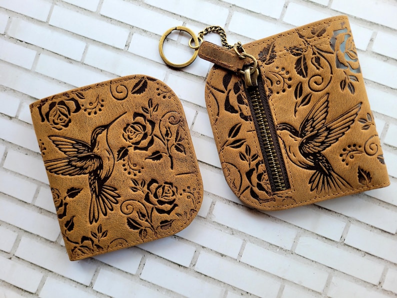Puede incluir: Dos carteras de cuero marr&oacute;n con dise&ntilde;os de colibr&iacute;es y rosas. Una cartera tiene cremallera y llavero. La otra es un tarjetero. Las carteras est&aacute;n grabadas con un estampado floral y un colibr&iacute;.