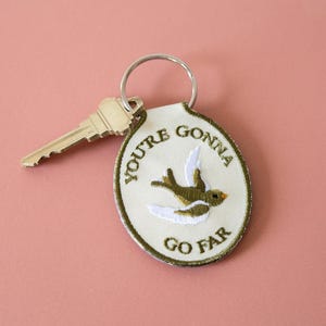 Può includere: Un portachiavi ovale color crema con la scritta ricamata "YOU'RE GONNA GO FAR" e un disegno di uccello. Il portachiavi ha un bordo verde e un portachiavi argentato. Una chiave d'argento è accanto al portachiavi.