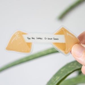 Puede incluir: Una pegatina transparente con forma de galleta de la fortuna. La pegatina tiene un diseño de galleta de color amarillo-naranja con el texto "You Are Capable Of Great Things" en una tira blanca.