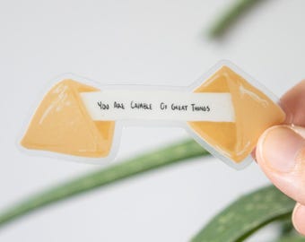 Puede incluir: Una pegatina transparente con forma de galleta de la fortuna. La pegatina tiene un diseño de galleta de color amarillo-naranja con el texto "You Are Capable Of Great Things" en una tira blanca.