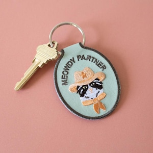 Könnte beinhalten: Ein Schlüsselanhänger mit einem hellgrünen ovalen Anhänger mit einer Katze mit Cowboyhut und den Worten "MEOWDY PARTNER". Ein goldfarbener Schlüssel ist an dem silbernen Schlüsselring befestigt. Der Hintergrund ist einfarbig rosa.