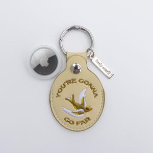 Può includere: Un portachiavi in pelle beige con le parole ricamate "YOU'RE GONNA GO FAR" e un disegno di uccello. Include un portachiavi argentato, una piccola targhetta in metallo e un AirTag Apple bianco e nero.