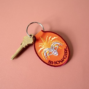 Könnte beinhalten: Orangefarbener, bestickter Schlüsselanhänger mit Spinnendesign und dem Wort "SHOWGIRL" in einem Kreis. Ein goldfarbener Schlüssel ist am Schlüsselring befestigt. Der Hintergrund ist einfarbiges, gedämpftes Rosa.