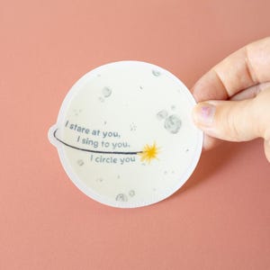 May include: A circular sticker featuring a pale off-white background with grey craters and a yellow star. A black curved line orbits the star. The text reads: I stare at you, I sing to you, I circle you. The sticker has a small tab on the left side.