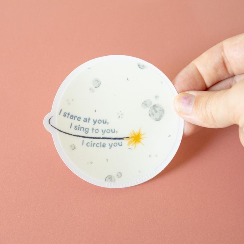 May include: A circular sticker featuring a pale off-white background with grey craters and a yellow star. A black curved line orbits the star. The text reads: I stare at you, I sing to you, I circle you. The sticker has a small tab on the left side.
