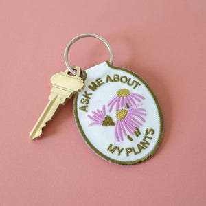 May include: A white oval-shaped keychain with green embroidered text that reads "Ask Me About My Plants". The keychain features two pink embroidered flowers with yellow centers. A gold key is attached to the keychain.