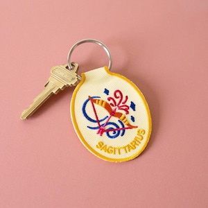 May include: An oval-shaped cream-colored embroidered keychain featuring a red and orange bow and arrow design with blue accents and the text SAGITTARIUS stitched in yellow. A silver metal split ring holds the keychain and a brass-colored key.