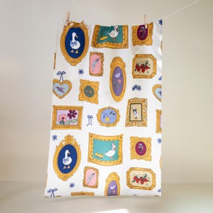 May include: A white tea towel with a repeating pattern of gold picture frames. Each frame contains a different image, including a goose, flowers, a lemon, and a bird. The background is white with small blue flowers.