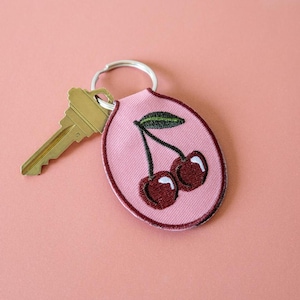 Op de afbeelding: Een roze stoffen sleutelhanger met een kersendesign. De sleutelhanger heeft een geborduurd beeld van twee kersen met een groen blad en een donkerrode omtrek. Een goudkleurige sleutel is aan de sleutelhanger bevestigd.