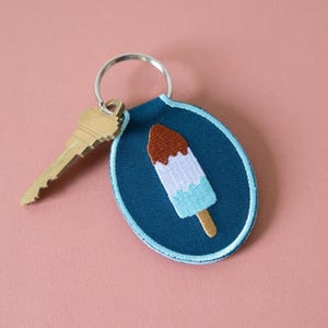 May include: A teal keychain with a popsicle design. The popsicle has a brown top, white center, and light blue bottom, with a wooden stick. A gold key is attached to the keychain.