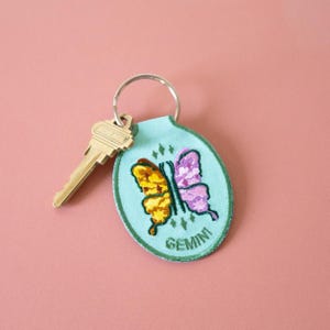 May include: A light blue oval keychain featuring an embroidered butterfly with one yellow wing and one purple wing. The word GEMINI is stitched in green at the bottom. A silver metal key ring is attached to the top, holding a brass-colored key.