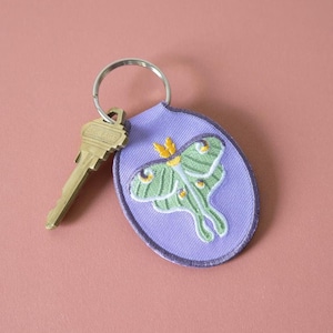 May include: A key ring with a lavender oval tag featuring an embroidered green and white moth design. A gold key is attached to the ring. The moth has yellow accents. The key ring is a practical accessory.