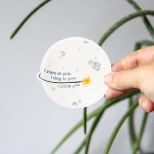 May include: A circular white sticker featuring a gray moon surface design with craters. The text reads, I stare at you, I sing to you, I circle you. A thin black line orbits the text, ending in a small yellow starburst graphic on the right side.