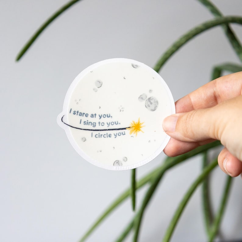 May include: A circular white sticker featuring a grey moon surface design with craters. The text reads, I stare at you, I sing to you, I circle you. A thin black line orbits the text, ending in a small yellow starburst graphic on the right side.