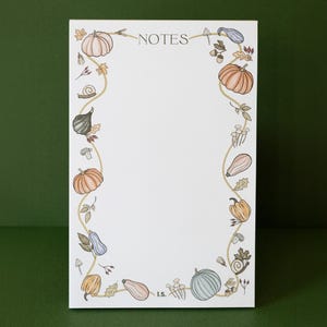 以下が含まれることがあります： かぼちゃ、かぼちゃ、葉っぱの装飾的な縁取りが施された白いメモ帳。メモ帳の上部には「NOTES」という文字が印刷され、下部には「I.S.」のイニシャルが印刷されています。