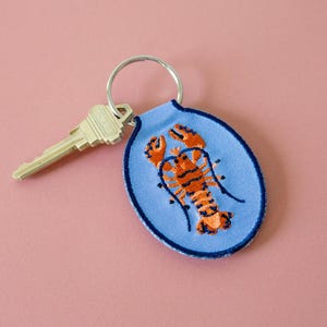 May include: A light blue keychain with a silver key ring and a key. The keychain features an embroidered orange and black lobster design within a blue oval shape. The key is silver.