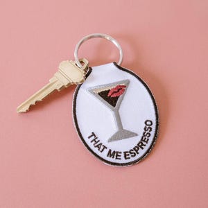 May include: White oval keychain with a martini glass design and the text "THAT ME ESPRESSO". The martini glass has a red lip design. A gold key is attached to the keychain ring. The background is a solid pink colour.