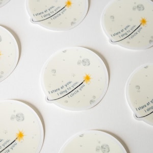 May include: Circular stickers featuring a pale moon illustration with grey craters. Each sticker displays a yellow star-tipped wand and the text: I stare at you, I sing to you, I circle you. The design includes a thin black orbital line.