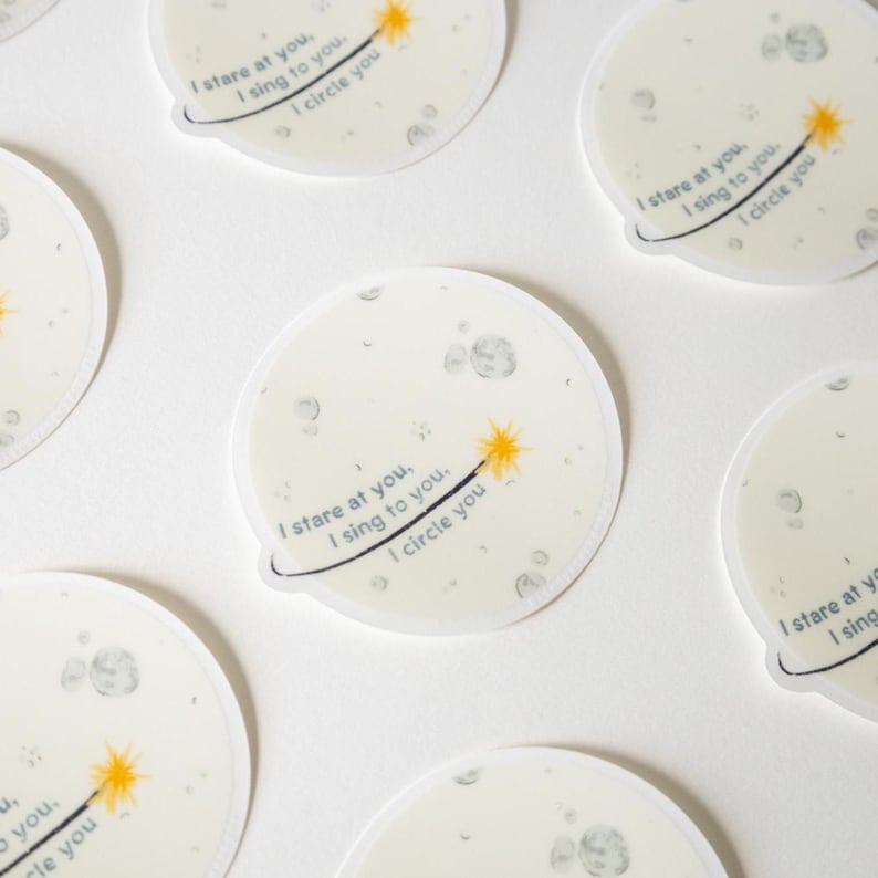 May include: Circular stickers featuring a pale moon illustration with grey craters. Each sticker displays a yellow star-tipped wand and the text: I stare at you, I sing to you, I circle you. The design includes a thin black orbital line.