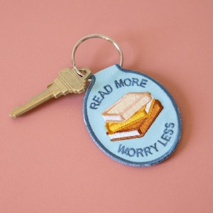Może przedstawiać: Jasnoniebieski brelok do kluczy ze srebrnym kółkiem i złotym kluczem. Brelok posiada haftowaną naszywkę z dwoma ułożonymi w stos książkami i napisem "Read More Worry Less" w granatowym kolorze.