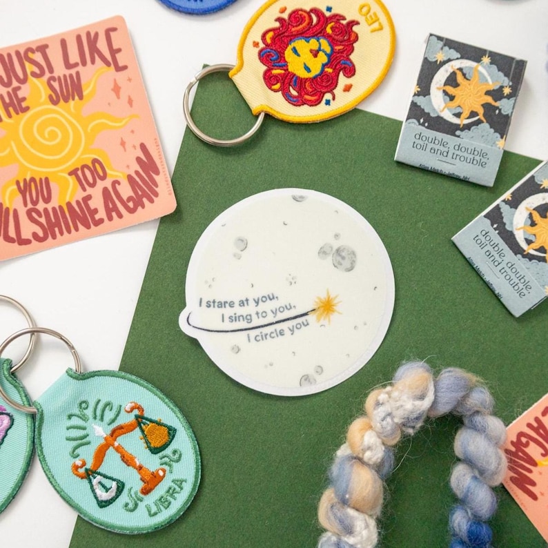 May include: A collection of items including a square card with a sun illustration, a round Leo keychain, two rectangular cards with moon graphics, a round sticker with moon art and text, a Libra keychain, and a braided blue and cream yarn strand.