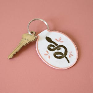Può includere: Un portachiavi ovale bianco con un design a serpente ricamato marrone e cuciture rosa. Il portachiavi ha un anello portachiavi argentato.