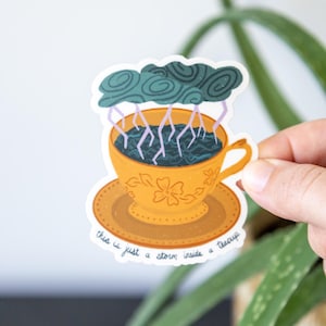 May include: A sticker depicting a yellow teacup and saucer, filled with dark blue liquid and lightning. Above the cup is a teal cloud. The text on the sticker reads, "this is just a storm inside a teacup."