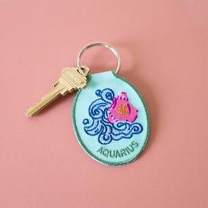 May include: A light blue oval keychain featuring an embroidered design of swirling dark blue water and a pink vessel with a yellow star. The word AQUARIUS is stitched in green at the bottom. A silver metal key ring holds a brass-colored key.