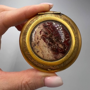 May include: A round, gold-coloured compact with a hinged lid. The compact has a decorative border and a red and white marbled design on the inside.