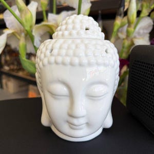 Puede incluir: Una estatua de cabeza de Buda de cerámica blanca con los ojos cerrados y una expresión serena. La cabeza tiene una superficie detallada y texturizada y una parte superior pequeña y redondeada. Ideal para la decoración del hogar.