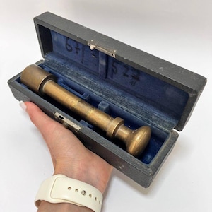 May include: A brass instrument, possibly a measuring tool, rests in a dark gray case with a blue velvet interior. The case is open, revealing the instrument's cylindrical shape and rounded end. The case is held in a hand.