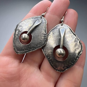 May include: A pair of silver earrings with a unique geometric design. Each earring features a textured, irregular-shaped metal piece with a circular cutout and a small ball hanging from a metal rod.