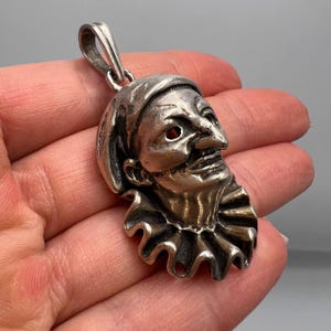 May include: Silver pendant featuring a detailed, three-dimensional sculpture of a jester's face with a ruffled collar.