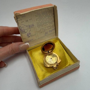 May include: A vintage gold-toned pocket watch with a hinged cover, displayed in its original box. The watch has a round face with visible numbers and hands. The box is lined with yellow fabric and includes a faded paper with text.