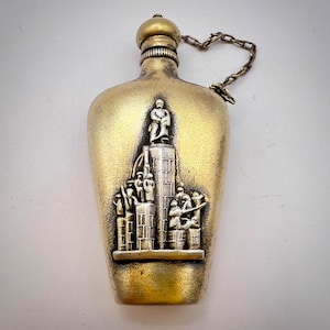 Puede incluir: Una petaca de metal de color dorado con un diseño en relieve plateado que representa a un grupo de personas de pie en una plataforma. La petaca tiene una cadena unida a la parte superior.