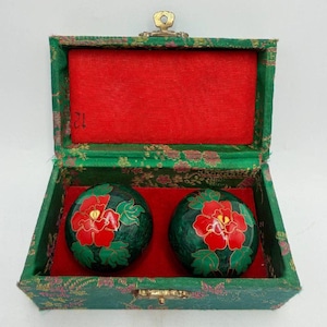 May include: Two green marble Baoding balls with red floral designs, displayed in an open green box with a red velvet interior. The box has a gold clasp and hinge.