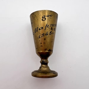 Puede incluir: Una pequeña copa de metal dorado con la parte superior acampanada y una base redonda. La copa tiene la inscripción "8 oz. Maftma 1968." en el lateral.