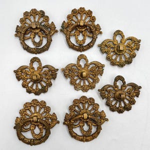 May include: Eight antique brass drawer pulls with detailed floral and scrollwork. Each pull has a ring handle and decorative backplate. The brass has an aged patina, giving them a vintage appearance. Ideal for furniture restoration.