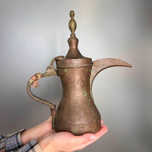 May include: Antique copper coffee pot with a long spout and ornate handle. The pot has a bulbous body, a lid, and a decorative finial. The copper has a warm, aged patina with hints of green oxidation. A vintage piece.