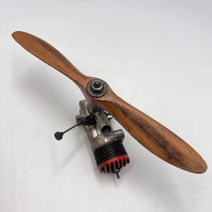 May include: A vintage model airplane engine with a wooden propeller. The propeller is a light brown colour and is attached to a silver-coloured engine with a red and black base. The engine has a small black knob and wire attached.