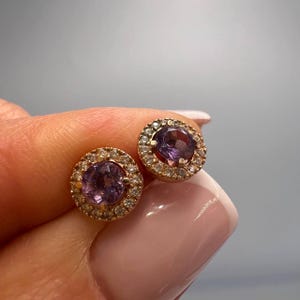 May include: A pair of gold stud earrings with a round purple gemstone surrounded by a halo of smaller clear gemstones.